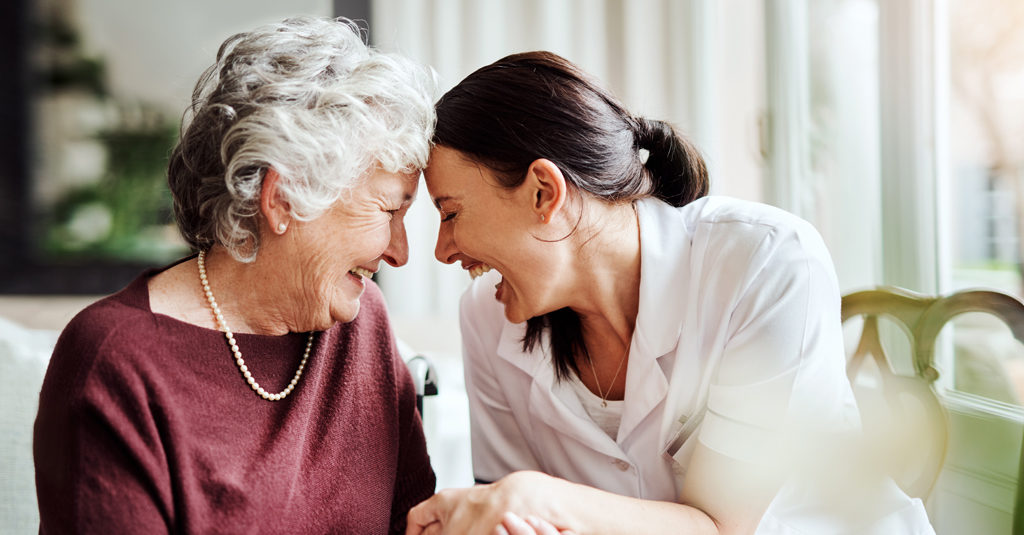 Two women leaning toward each other laughing. One is a nurse and the other is an elderly woman.