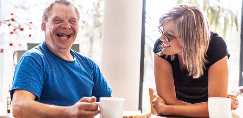 Happy man with a developmental disability sitting at a kitchen table with a smiling woman who is looking at him.