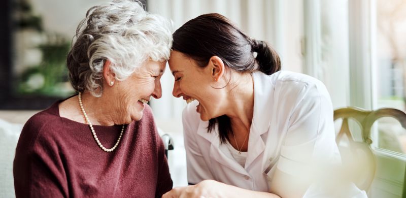 Two women leaning toward each other laughing. One is a nurse and the other is an elderly woman.